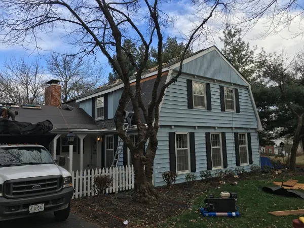 Blue two-story house undergoing roof work with ladder, pickup truck, white picket fence and a worker near leafless tree