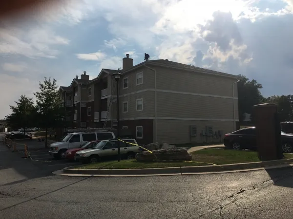 Apartment building with a person on the roof by a ladder, parked cars and caution tape in foreground under cloudy sky