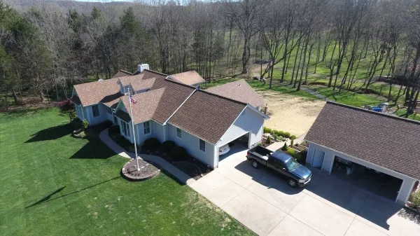 Aerial view of a single-story suburban home with detached garage, pickup truck, flagpole, driveway and surrounding trees.