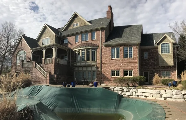 Large brick mansion with covered swimming pool, stone retaining wall and dry winter landscaping under a cloudy sky