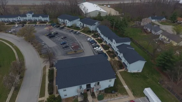 Aerial view of suburban apartment townhomes with parking lot, curved road, lawns and nearby trees.