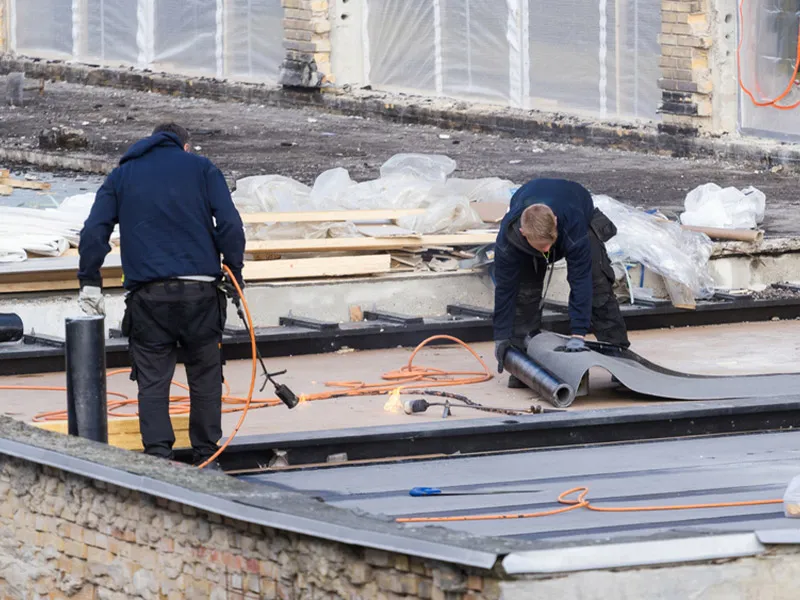 Two workers installing rolled roofing membrane on a flat roof using torches and tools