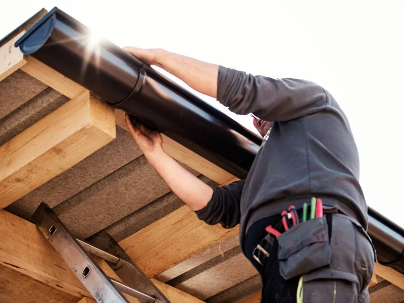 A person installing a black rain gutter on a wooden roof edge while standing on a ladder with a tool belt