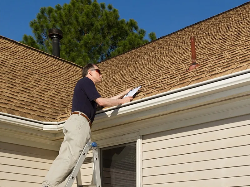 A man on a ladder inspects a shingled roof and takes notes beside the gutter under a clear blue sky