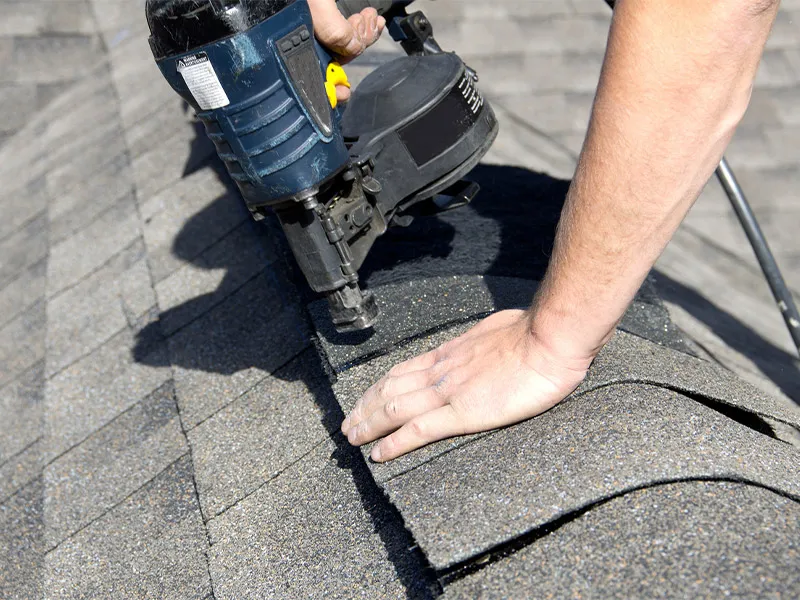 A person using a pneumatic nail gun to secure asphalt ridge cap shingles on a roof