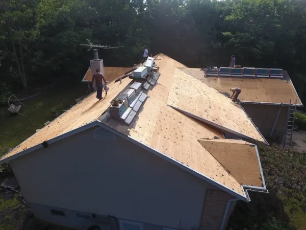 Workers installing plywood sheathing on a residential roof with stacked materials, tools and ladders